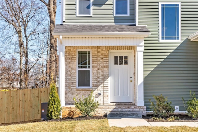 doorway to property with brick siding, fence, and roof with shingles