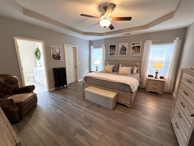 bedroom with ceiling fan, ensuite bathroom, a tray ceiling, and dark hardwood / wood-style flooring