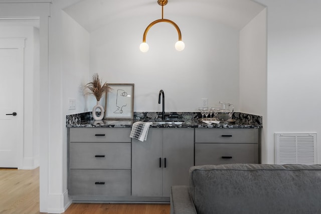 bar featuring sink, vaulted ceiling, light hardwood / wood-style flooring, dark stone countertops, and decorative light fixtures