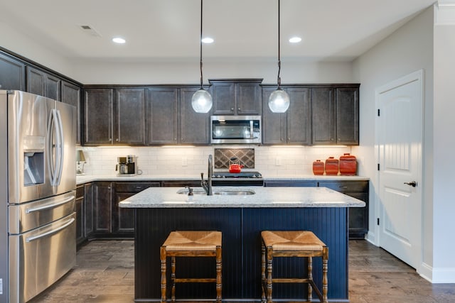 kitchen featuring light stone counters, dark hardwood / wood-style floors, an island with sink, pendant lighting, and appliances with stainless steel finishes