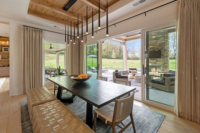 dining room featuring beam ceiling, ceiling fan, wooden ceiling, a stone fireplace, and light hardwood / wood-style floors