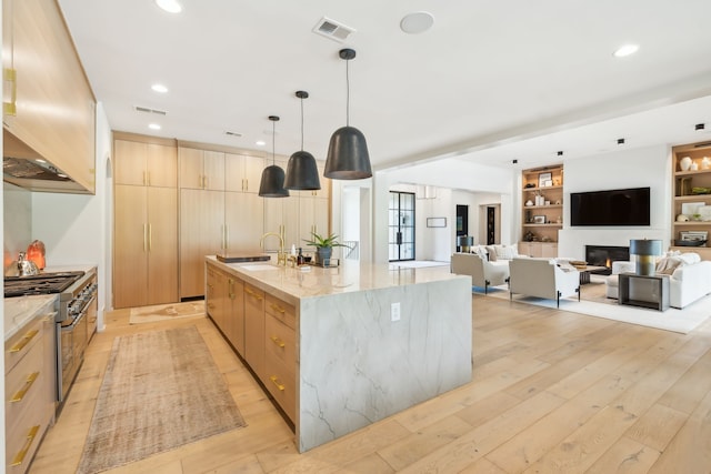 kitchen with light stone counters, a large island with sink, light hardwood / wood-style floors, light brown cabinetry, and stainless steel stove