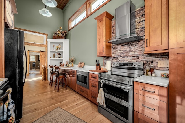 kitchen featuring wall chimney exhaust hood, hanging light fixtures, light hardwood / wood-style flooring, backsplash, and appliances with stainless steel finishes