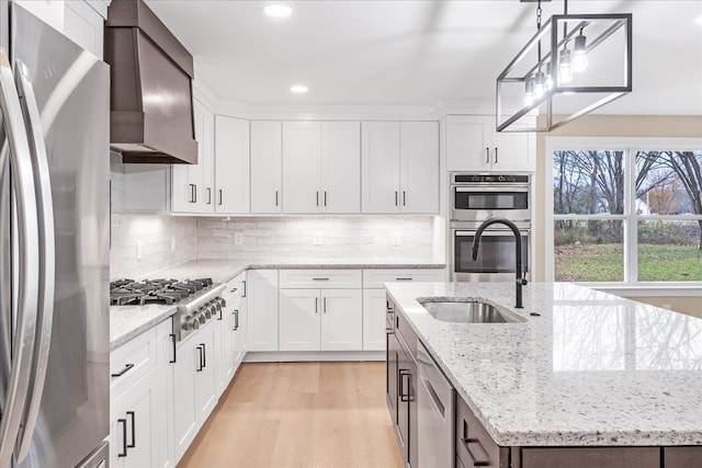 kitchen featuring light wood-type flooring, stainless steel appliances, wall chimney range hood, decorative light fixtures, and white cabinets