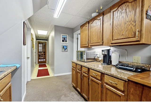 kitchen featuring brown cabinets, light colored carpet, stainless steel microwave, a drop ceiling, and baseboards