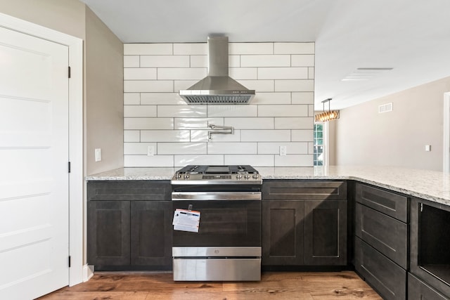 kitchen with backsplash, light stone counters, wall chimney range hood, wood-type flooring, and stainless steel stove