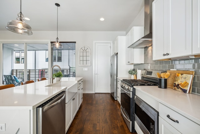 kitchen with appliances with stainless steel finishes, dark hardwood / wood-style flooring, wall chimney range hood, hanging light fixtures, and an island with sink