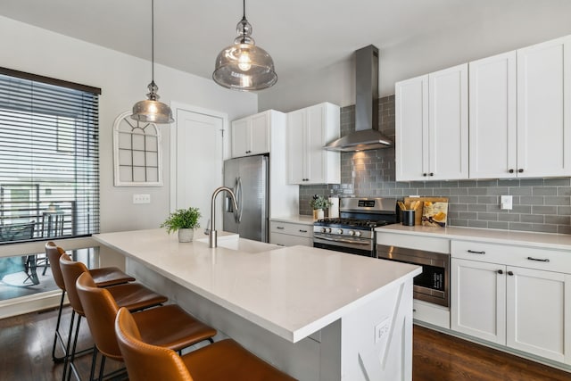 kitchen featuring wall chimney range hood, dark hardwood / wood-style floors, pendant lighting, a center island with sink, and appliances with stainless steel finishes