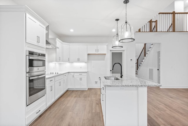 kitchen featuring sink, a kitchen island with sink, white cabinets, decorative light fixtures, and stainless steel double oven