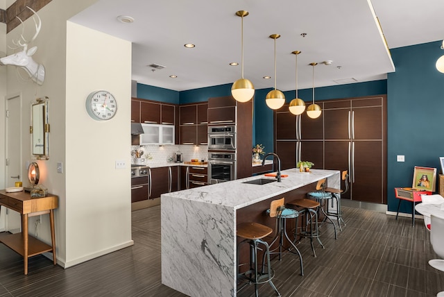 kitchen featuring a center island with sink, sink, decorative light fixtures, light stone counters, and dark brown cabinetry