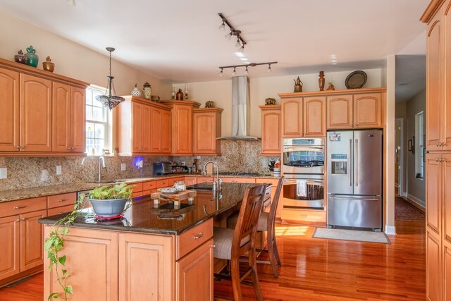 kitchen featuring appliances with stainless steel finishes, wall chimney exhaust hood, sink, light hardwood / wood-style floors, and an island with sink