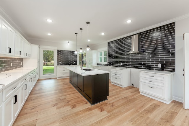 kitchen with light wood-type flooring, wall chimney exhaust hood, a kitchen island with sink, sink, and white cabinets