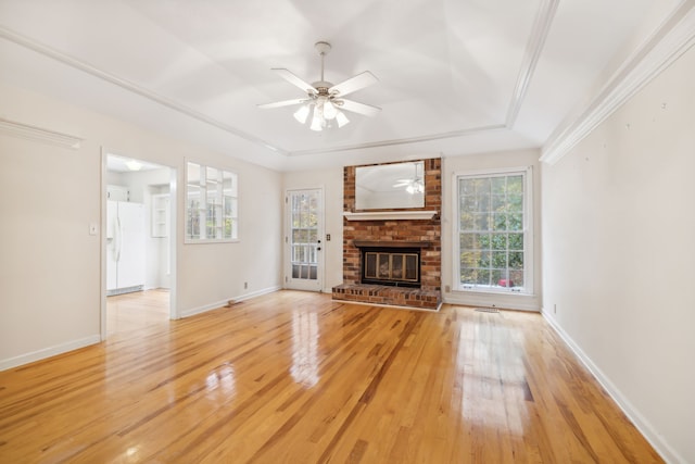 unfurnished living room featuring a brick fireplace, ornamental molding, a raised ceiling, ceiling fan, and light hardwood / wood-style floors