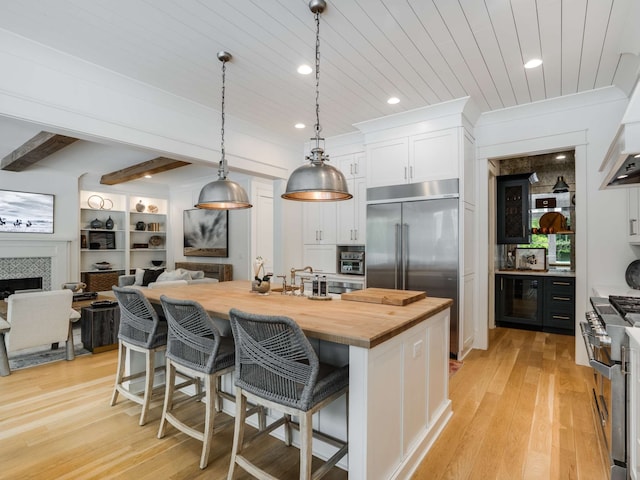 kitchen featuring a kitchen island with sink, a fireplace, white cabinetry, hanging light fixtures, and butcher block counters