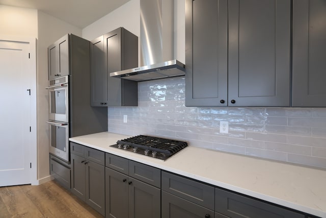 kitchen featuring appliances with stainless steel finishes, tasteful backsplash, wall chimney exhaust hood, gray cabinetry, and light hardwood / wood-style floors