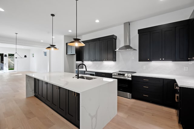 kitchen featuring stainless steel electric range oven, sink, hanging light fixtures, wall chimney range hood, and a large island with sink