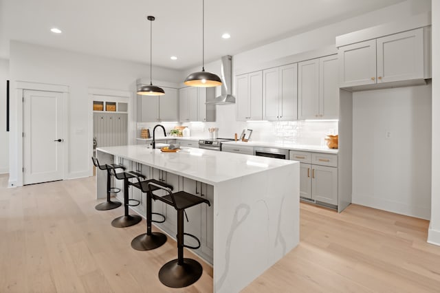 kitchen featuring a center island with sink, wall chimney range hood, decorative light fixtures, light hardwood / wood-style floors, and light stone counters