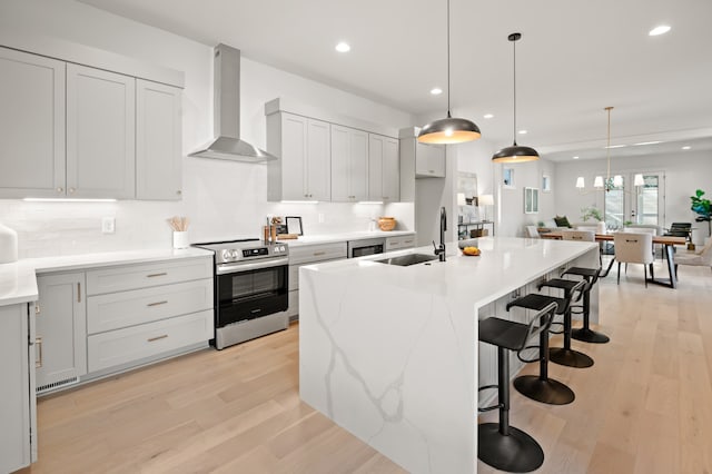 kitchen with wall chimney range hood, light hardwood / wood-style flooring, decorative light fixtures, electric stove, and a kitchen island with sink