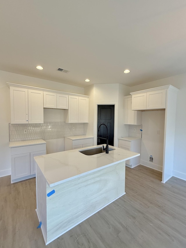 kitchen with backsplash, light hardwood / wood-style flooring, light stone countertops, an island with sink, and white cabinetry