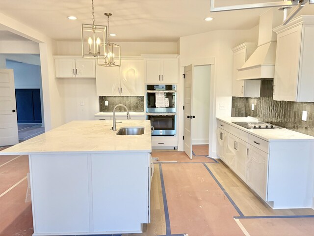 kitchen featuring custom range hood, hanging light fixtures, a kitchen island with sink, stainless steel double oven, and a sink