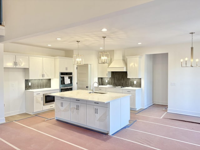 kitchen with a kitchen island with sink, premium range hood, a sink, and white cabinetry