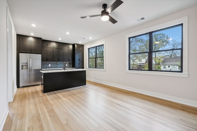 kitchen with decorative backsplash, stainless steel fridge, ceiling fan, a center island with sink, and light hardwood / wood-style floors