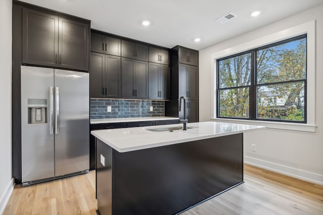 kitchen with stainless steel fridge, backsplash, dark brown cabinets, a kitchen island with sink, and light hardwood / wood-style flooring