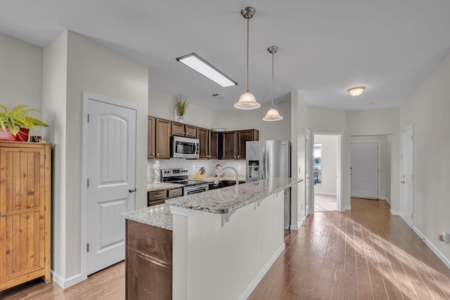 kitchen featuring hanging light fixtures, stainless steel appliances, tasteful backsplash, a kitchen island with sink, and light wood-type flooring