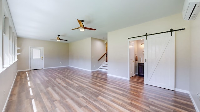 unfurnished living room featuring wood-type flooring, a barn door, a wall mounted AC, and ceiling fan