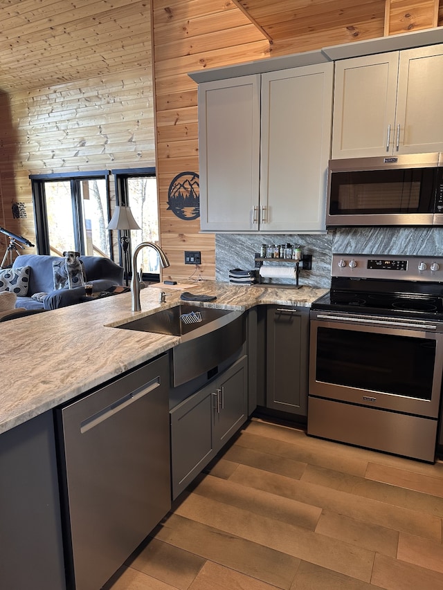 kitchen with light stone countertops, sink, stainless steel appliances, gray cabinets, and wooden walls