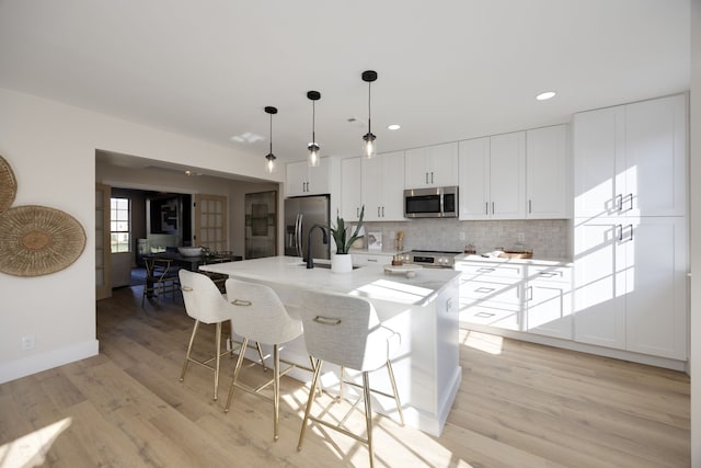 kitchen with light wood-type flooring, stainless steel appliances, decorative light fixtures, white cabinetry, and an island with sink