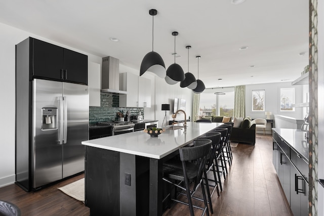 kitchen featuring appliances with stainless steel finishes, wall chimney exhaust hood, dark wood-type flooring, hanging light fixtures, and an island with sink