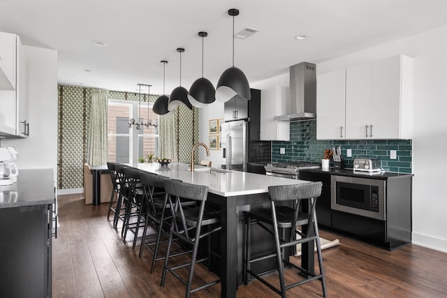 kitchen featuring stainless steel appliances, dark wood-type flooring, wall chimney range hood, white cabinetry, and an island with sink