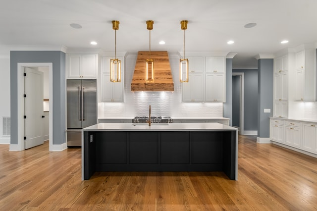 kitchen with hanging light fixtures, built in fridge, extractor fan, a kitchen island with sink, and light wood-type flooring