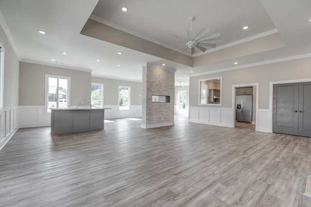 unfurnished living room with a raised ceiling, a stone fireplace, crown molding, ceiling fan, and light wood-type flooring