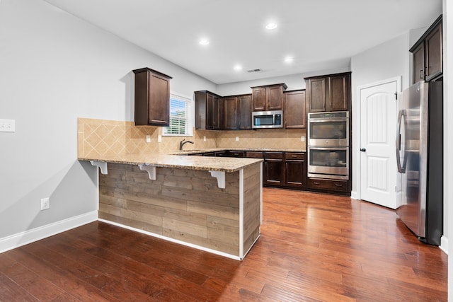 kitchen with kitchen peninsula, appliances with stainless steel finishes, a kitchen breakfast bar, light stone counters, and dark hardwood / wood-style floors