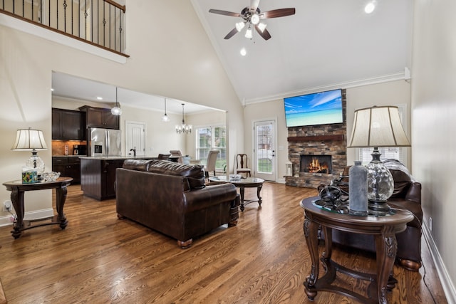 living room with a stone fireplace, high vaulted ceiling, dark hardwood / wood-style floors, crown molding, and ceiling fan with notable chandelier