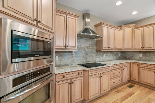 kitchen with light brown cabinetry, light wood-type flooring, backsplash, stainless steel appliances, and wall chimney range hood