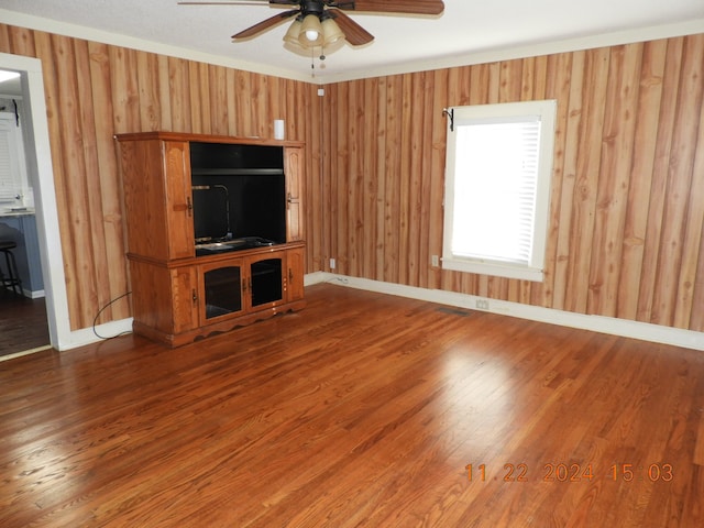 unfurnished living room with wood-type flooring, wooden walls, ceiling fan, and ornamental molding