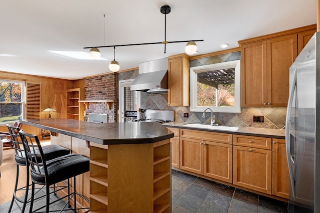 kitchen with wall chimney range hood, sink, stainless steel fridge, decorative light fixtures, and a kitchen island