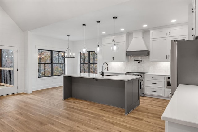 kitchen with light wood-type flooring, custom range hood, an island with sink, and appliances with stainless steel finishes