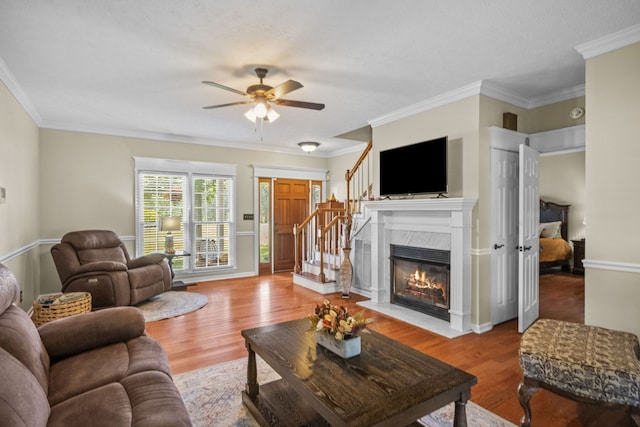 living room with ceiling fan, a fireplace with flush hearth, wood finished floors, stairs, and crown molding