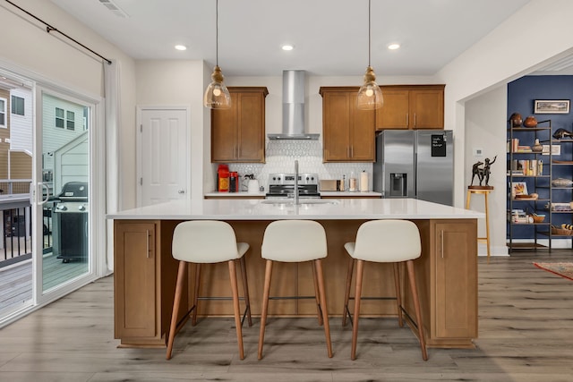 kitchen with stainless steel appliances, wall chimney range hood, pendant lighting, a kitchen island with sink, and light wood-type flooring