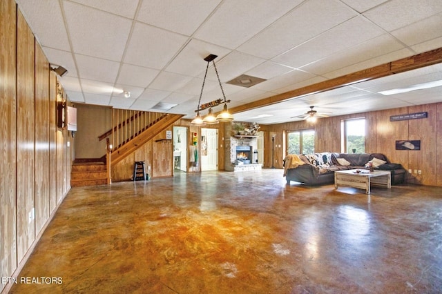 unfurnished living room featuring ceiling fan, a stone fireplace, wood walls, concrete flooring, and a paneled ceiling