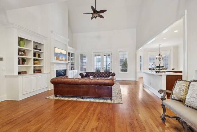 living room with french doors, built in shelves, high vaulted ceiling, light wood-type flooring, and ceiling fan with notable chandelier