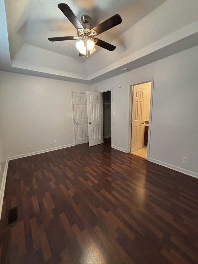 unfurnished bedroom with ensuite bath, ceiling fan, dark wood-type flooring, a textured ceiling, and a tray ceiling