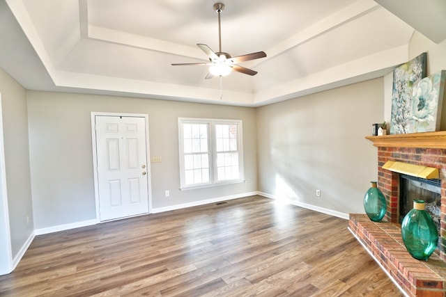 unfurnished living room with a raised ceiling, ceiling fan, wood-type flooring, and a brick fireplace