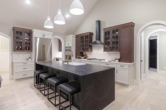 kitchen with sink, stainless steel appliances, wall chimney range hood, a kitchen island with sink, and white cabinets