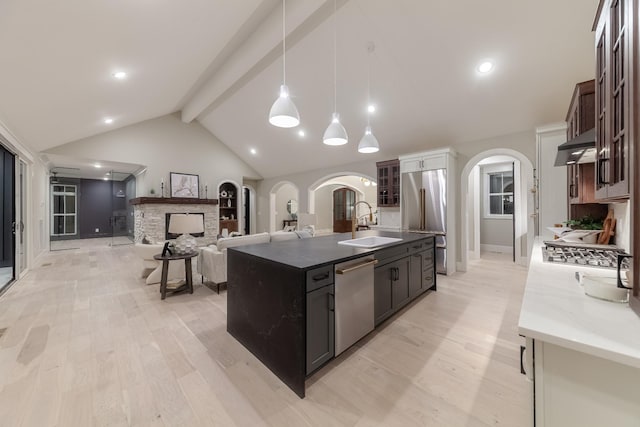 kitchen with light wood-type flooring, stainless steel appliances, a kitchen island with sink, vaulted ceiling with beams, and a stone fireplace