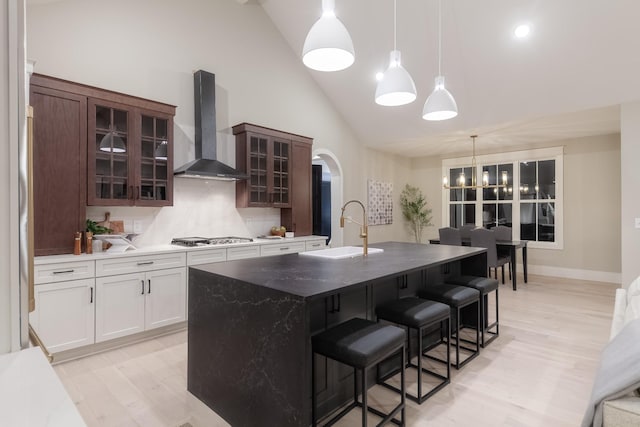 kitchen featuring dark brown cabinetry, sink, wall chimney exhaust hood, an island with sink, and light wood-type flooring